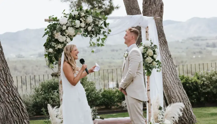 Novia ríe mientras lee sus votos, novio sonríe. Bodas sencillas pero bonitas, con arco floral y paisaje de fondo.