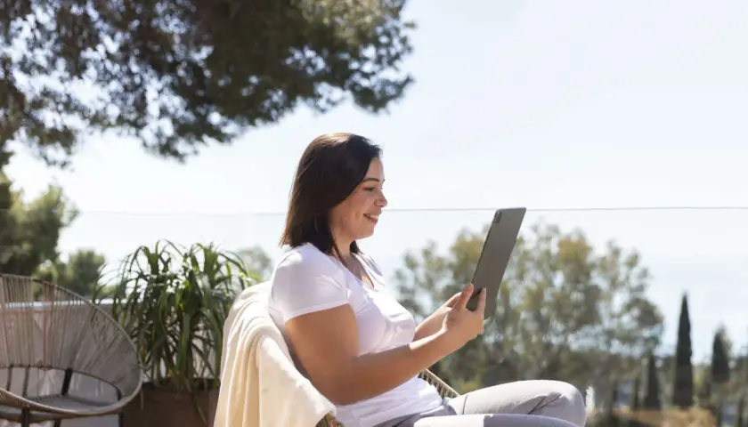 Mujer sonríe mientras trabaja en su tablet al aire libre, disfrutando de la vida de los trabajos para nómadas digitales.