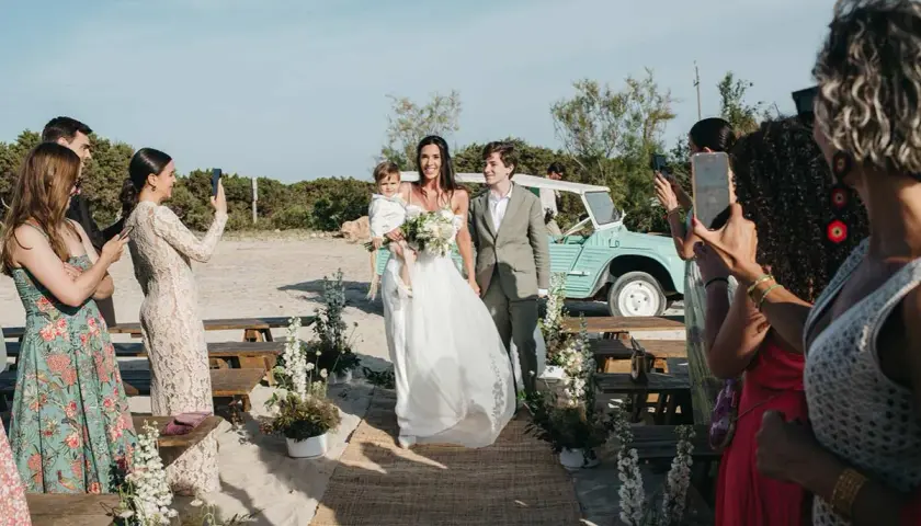 Novios caminan por la playa en una boda elegante. La novia lleva un niño en brazos. Un coche vintage espera.