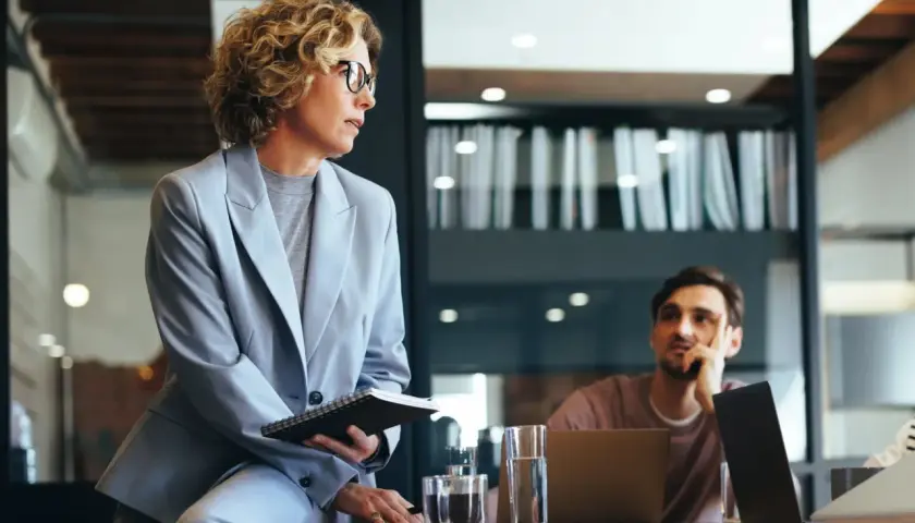 Mujer con gafas y traje gris, con cuaderno, en reunión. Podría ser parte de un curso organización de eventos gratis.