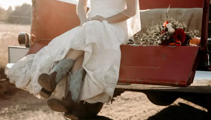 Novia con vestido de encaje y botas vaqueras, sentada en la parte trasera de una camioneta roja, con un ramo de flores. Un toque rústico para bodas vintage.