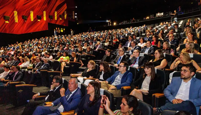 Amplia audiencia en un auditorio oscuro, asistiendo a eventos de empresa. El telón rojo brillante y las luces del escenario iluminan la sala llena de gente.