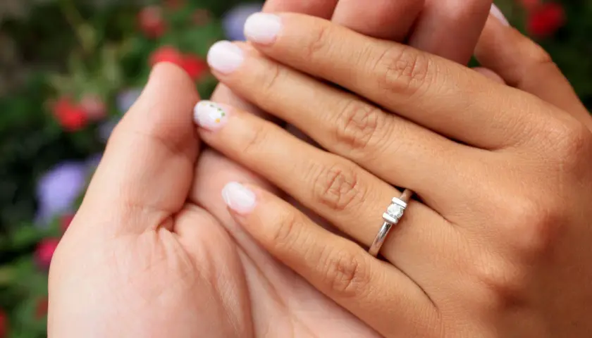 Manos entrelazadas, un dedo con un anillo de compromiso brilla entre flores.