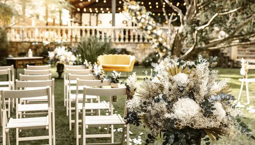Un arreglo floral blanco y dorado decora un jardín para una boda civil sencilla y barata. Sillas de madera esperan a los invitados.