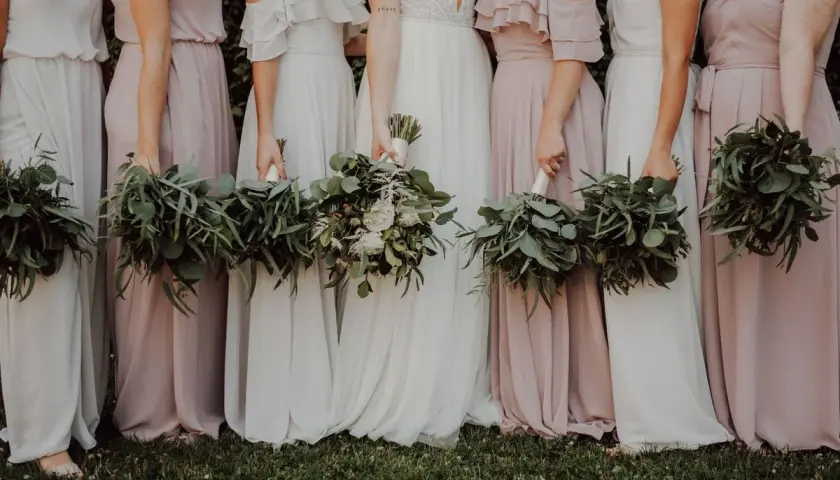 Un grupo de damas de honor en España, vestidas con trajes largos y fluidos en tonos pastel, sostienen ramos de eucalipto y flores blancas.