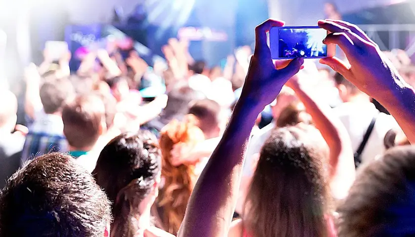 Manos sostienen un teléfono para grabar un evento comercial. La multitud levanta los brazos, disfrutando de la música y las luces.