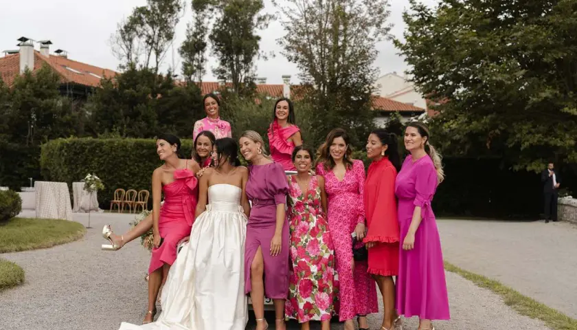 Grupo de mujeres sonriendo, vestidas con trajes rosas y morados. La novia, con un vestido blanco, está rodeada por su corte de honor, incluyendo a un hombre como dama de honor.
