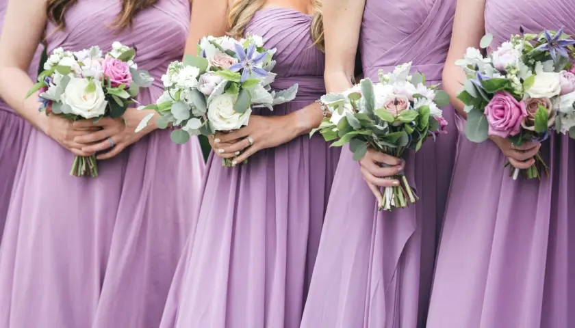 Damas de honor solteras, vestidas de lila, sostienen ramos de flores.