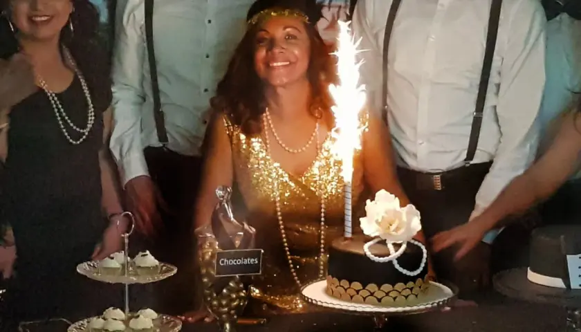 Una mujer sonriente con un vestido dorado y perlas celebra en una fiesta temática de los años 20. Hay un pastel con bengalas y dulces.