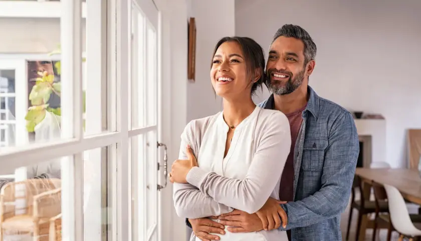 Pareja sonriente y abrazada, mirando por la ventana. Refleja cómo debe ser una relación de pareja: amor, apoyo y futuro compartido.