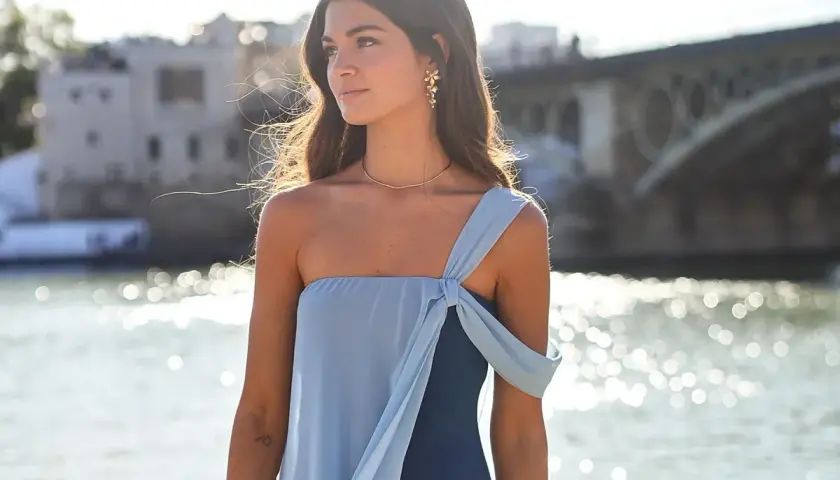 Mujer con vestido **boho chic** azul, con detalles de tela en el hombro, junto a un río con un puente al fondo.