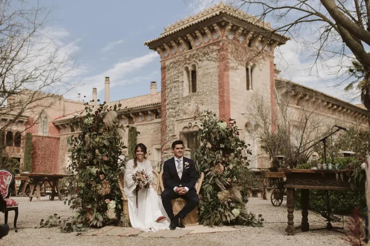 Novios sentados en sillas de mimbre, rodeados de exuberante vegetación y flores, con una arquitectura histórica de fondo. La decoración boda boho crea un ambiente romántico y natural.