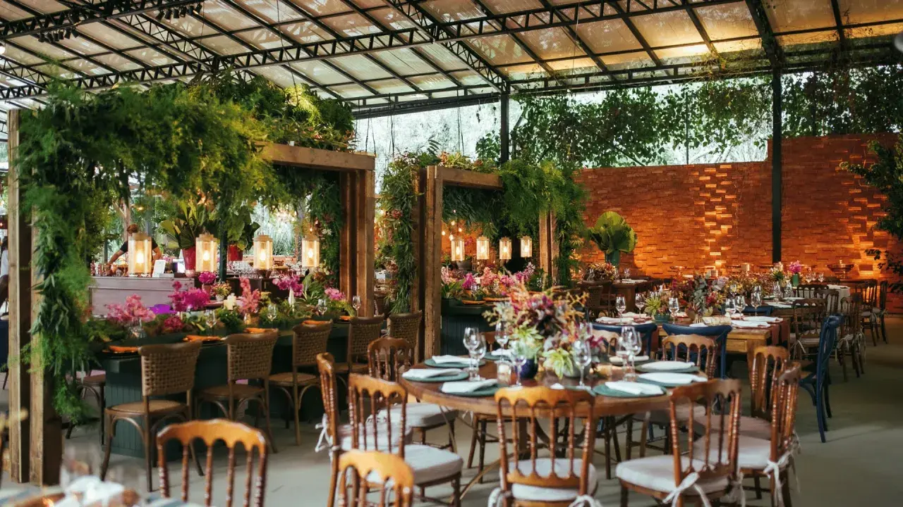 Salón de eventos decorado con abundante vegetación, flores y luces cálidas, con mesas puestas para una celebración.