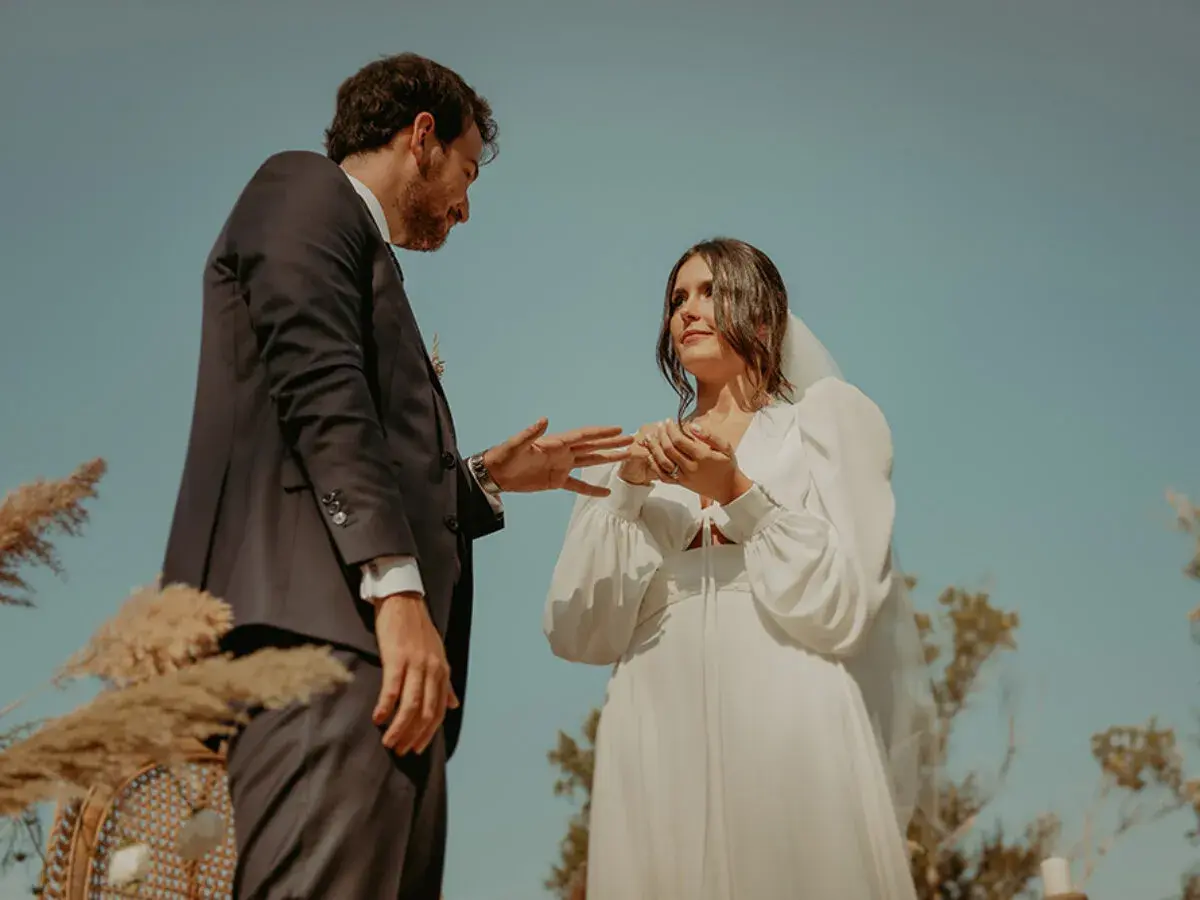 Novios intercambian anillos en una boda civil informal al aire libre. Un vestido blanco y un traje oscuro, rodeados de naturaleza.