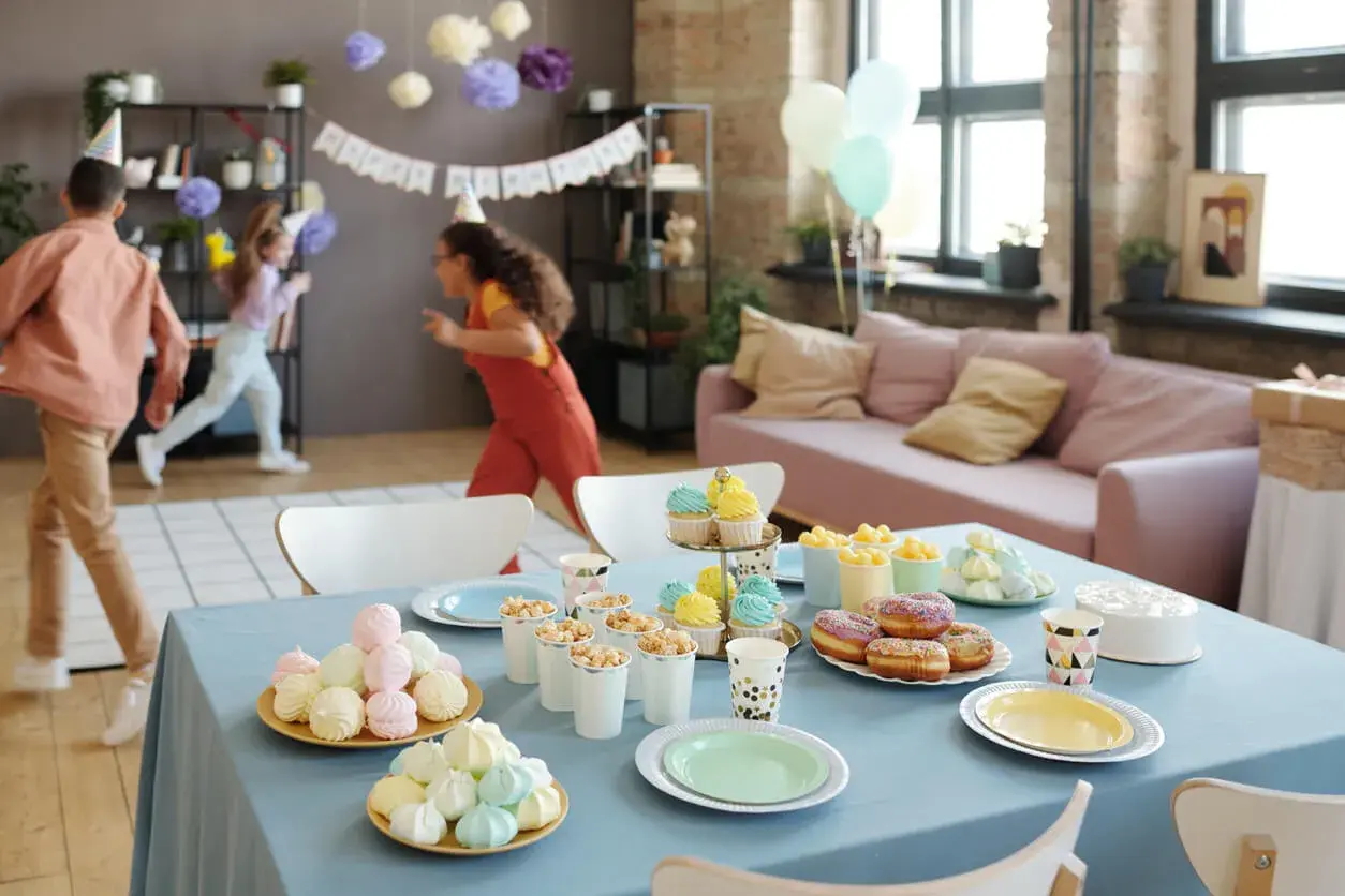 Niños corriendo en una fiesta de cumpleaños, con mesa llena de dulces y pastel. ¡Ideas de qué hacer en una fiesta!