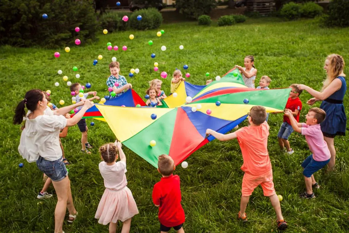 Niños y adultos juegan con un paracaídas de colores y pelotas, celebrando una fiesta de cumpleaños original en el césped.