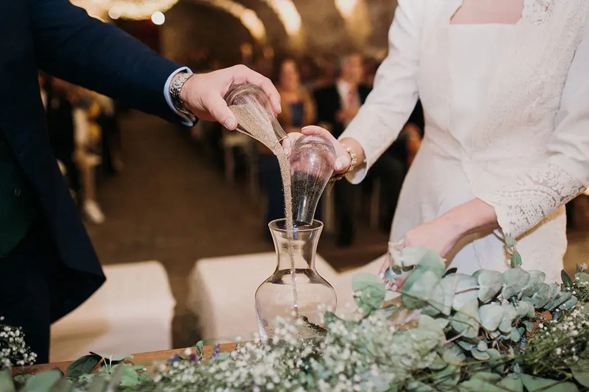 Manos de novios vierten arena en un jarrón, simbolizando su unión en una boda simbólica.