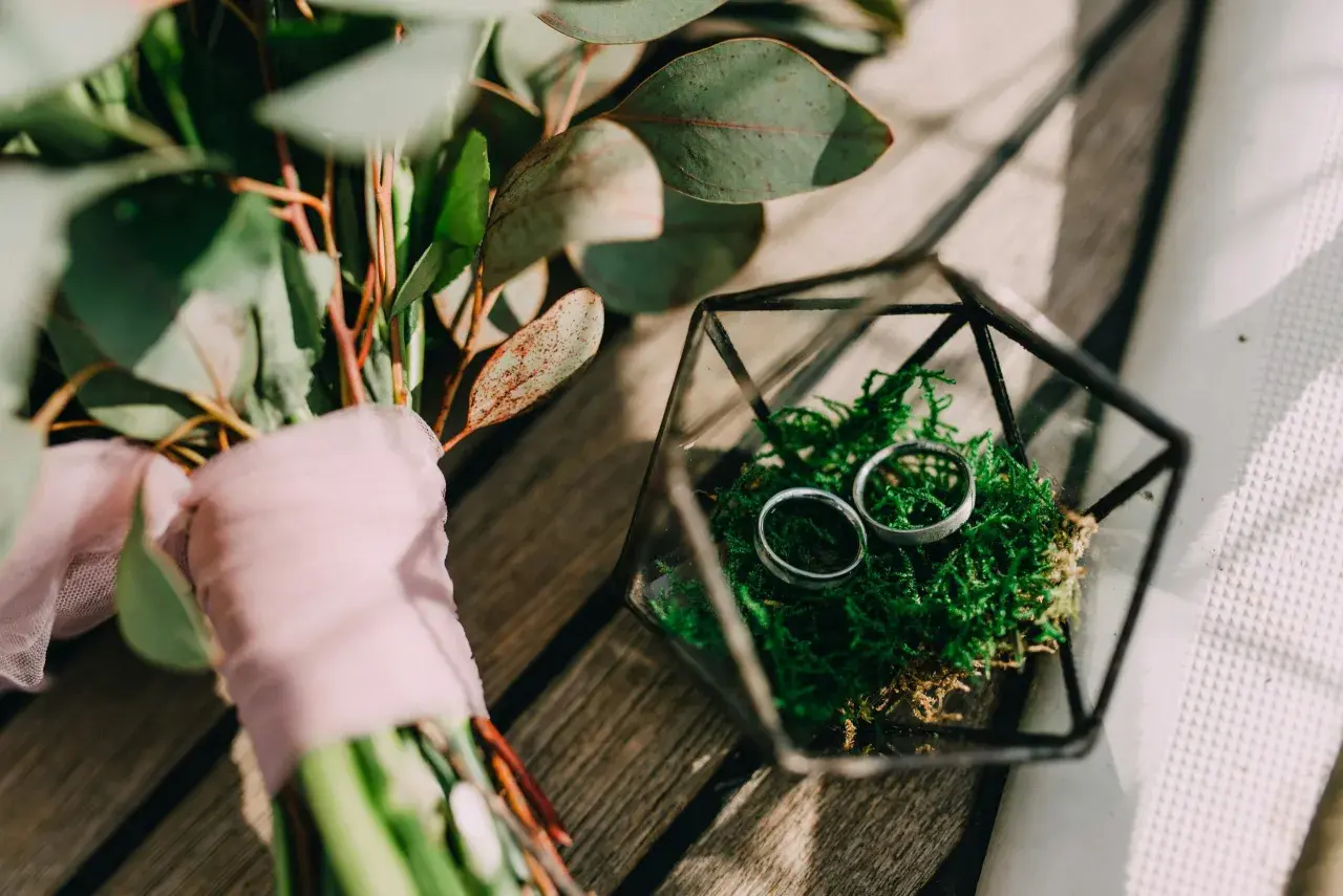 Anillos de boda en musgo verde dentro de un terrario de vidrio, junto a un ramo con tela rosa. Celebrando 5 años de casados.