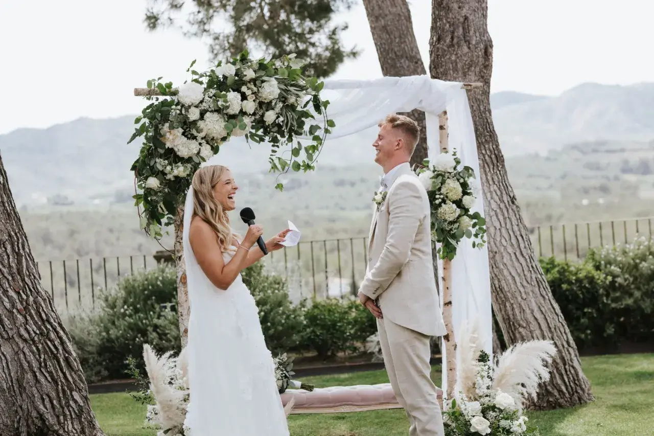 Novia ríe mientras lee sus votos, novio sonríe. Bodas sencillas pero bonitas, con arco floral y paisaje de fondo.