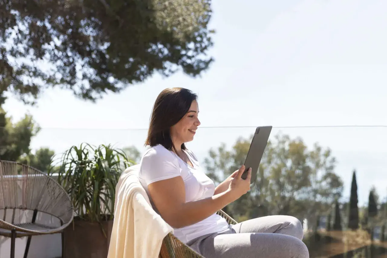 Mujer sonríe mientras trabaja en su tablet al aire libre, disfrutando de la vida de los trabajos para nómadas digitales.