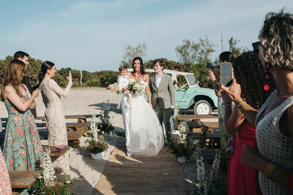 Novios caminan por la playa en una boda elegante. La novia lleva un niño en brazos. Un coche vintage espera.