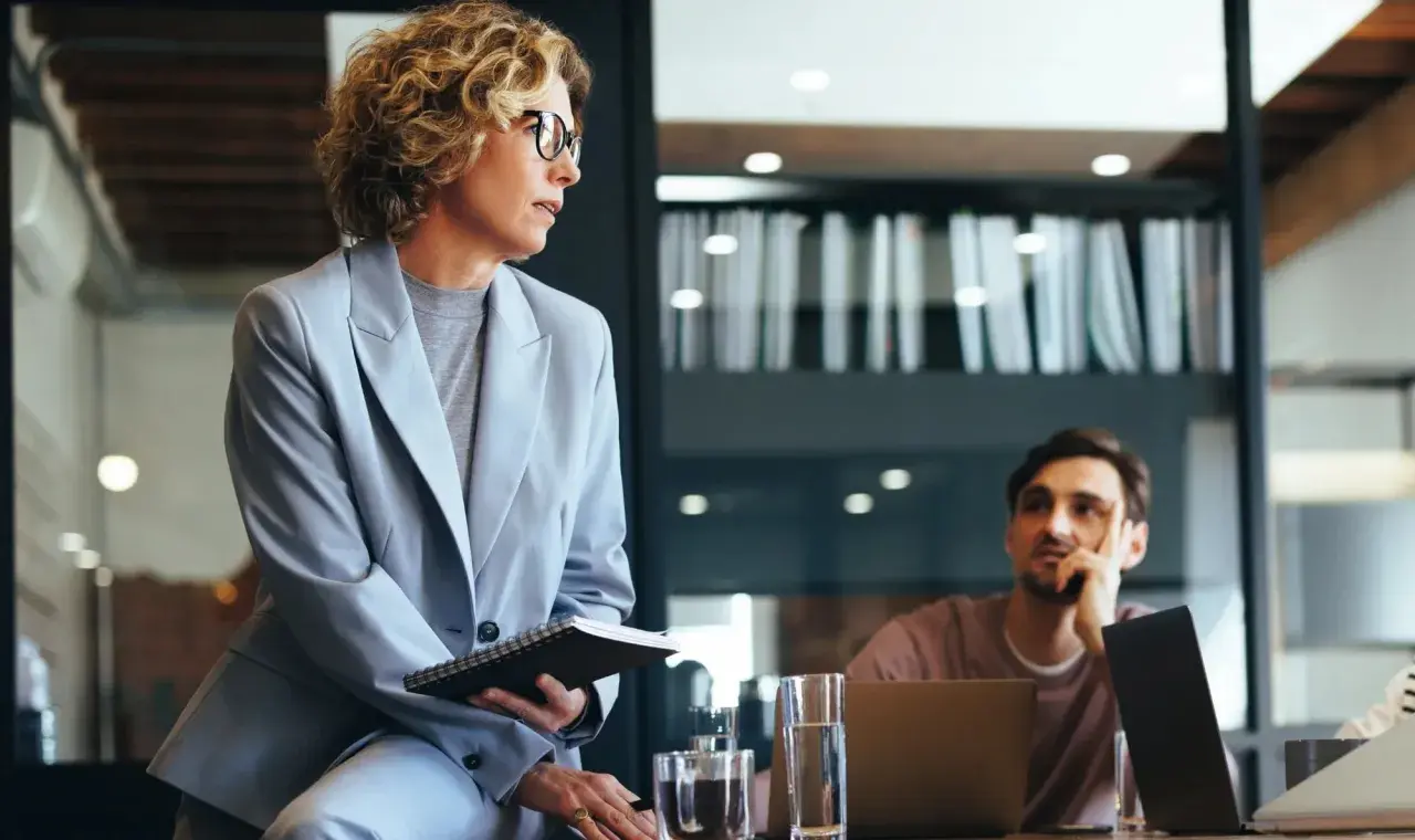 Mujer con gafas y traje gris, con cuaderno, en reunión. Podría ser parte de un curso organización de eventos gratis.