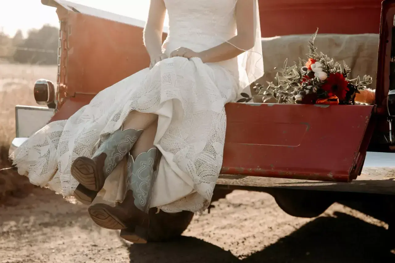 Novia con vestido de encaje y botas vaqueras, sentada en la parte trasera de una camioneta roja, con un ramo de flores. Un toque rústico para bodas vintage.