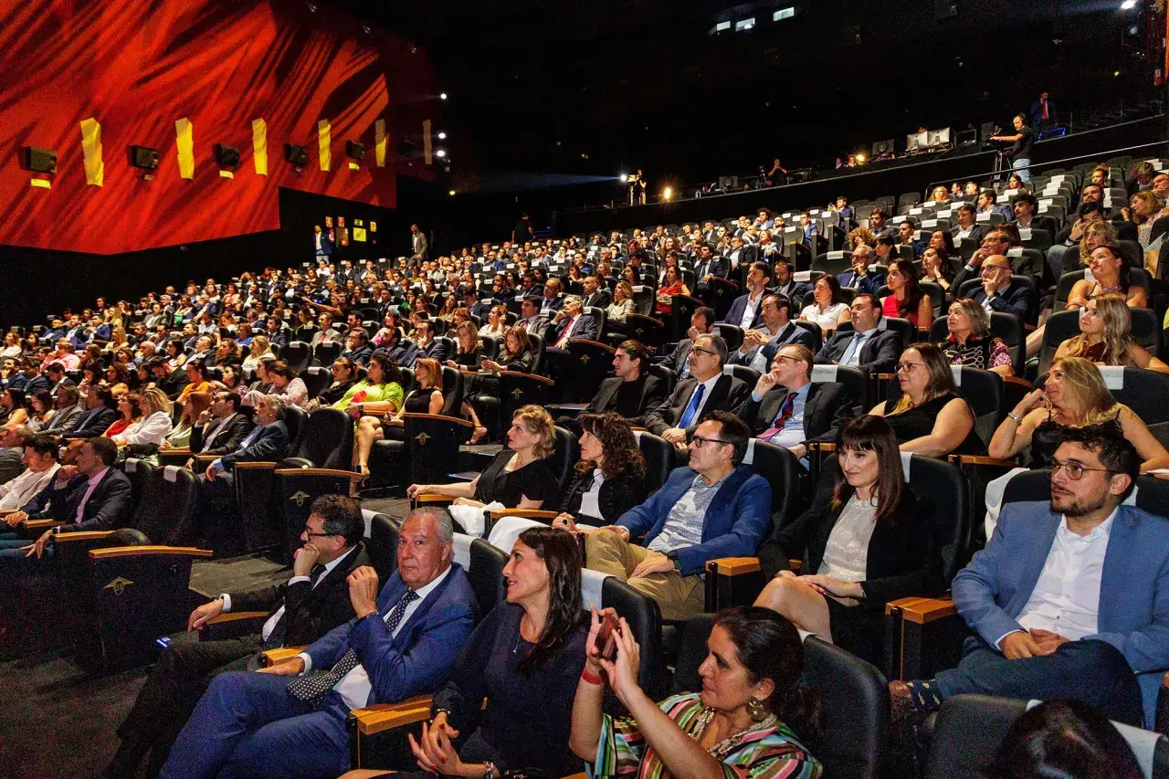 Amplia audiencia en un auditorio oscuro, asistiendo a eventos de empresa. El telón rojo brillante y las luces del escenario iluminan la sala llena de gente.