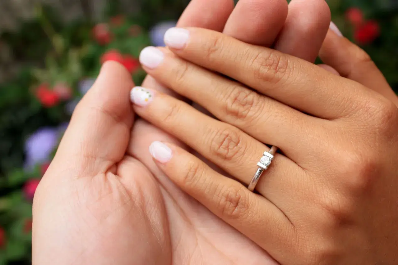 Manos entrelazadas, un dedo con un anillo de compromiso brilla entre flores.