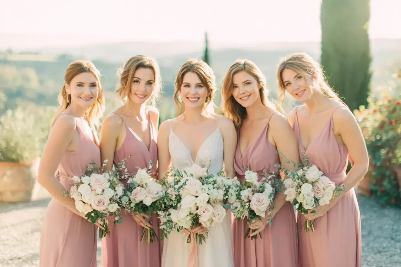 Novia sonriente con su damo de honor, todas con vestidos rosas y ramos de flores.