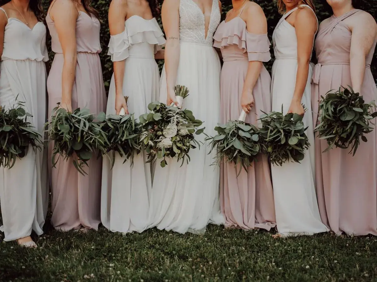 Un grupo de damas de honor en España, vestidas con trajes largos y fluidos en tonos pastel, sostienen ramos de eucalipto y flores blancas.