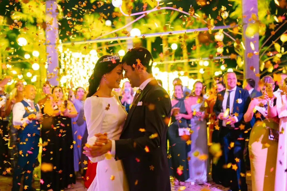 Una pareja baila felizmente en su **noche de bodas**, rodeada de confeti dorado y luces cálidas. Invitados sonríen al fondo.