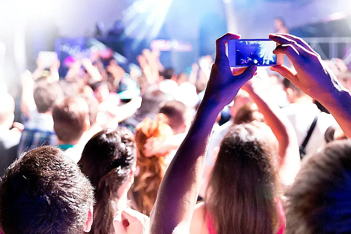 Manos sostienen un teléfono para grabar un evento comercial. La multitud levanta los brazos, disfrutando de la música y las luces.