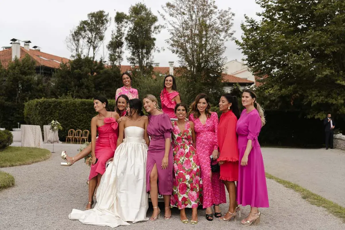 Grupo de mujeres sonriendo, vestidas con trajes rosas y morados. La novia, con un vestido blanco, está rodeada por su corte de honor, incluyendo a un hombre como dama de honor.