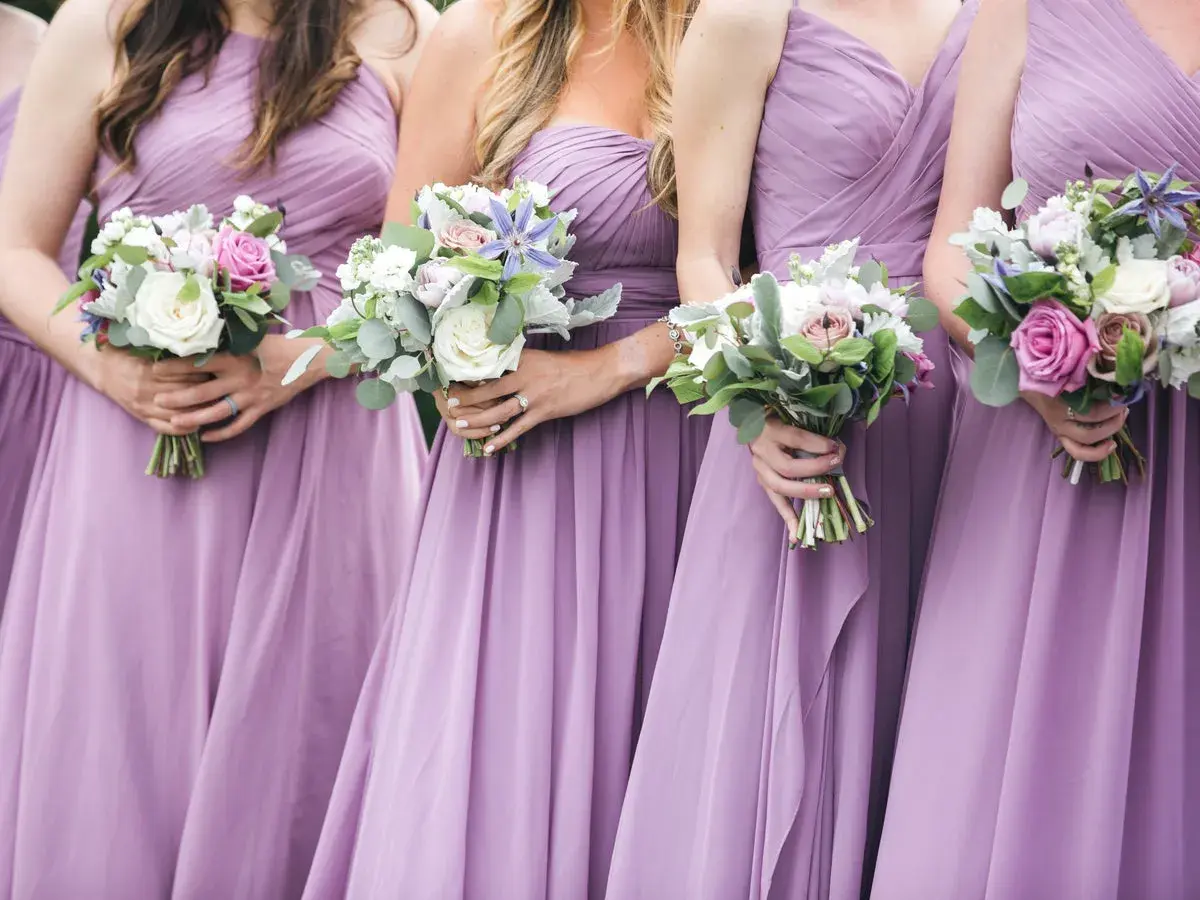 Damas de honor solteras, vestidas de lila, sostienen ramos de flores.