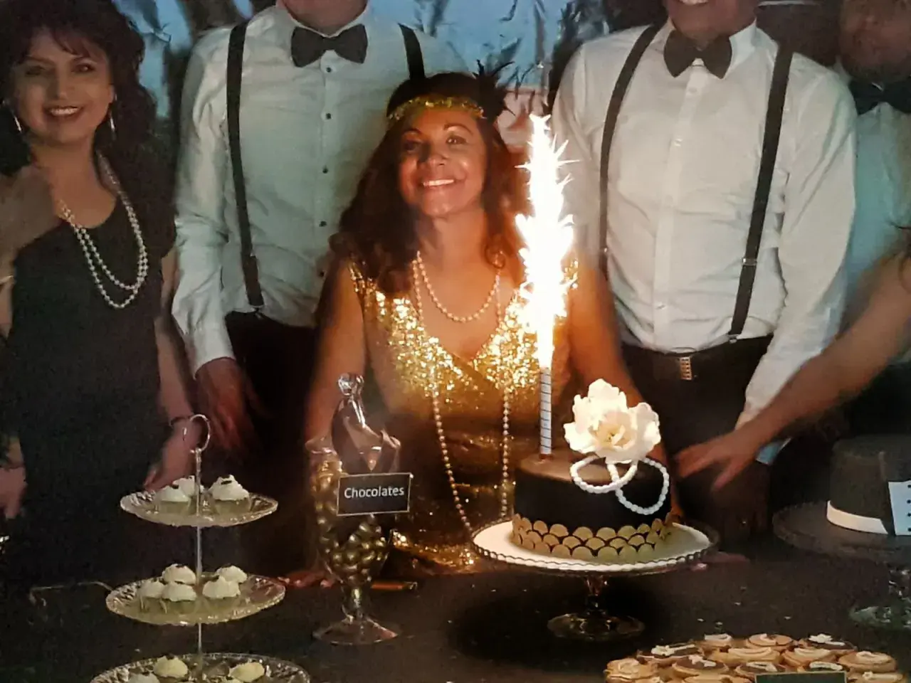 Una mujer sonriente con un vestido dorado y perlas celebra en una fiesta temática de los años 20. Hay un pastel con bengalas y dulces.