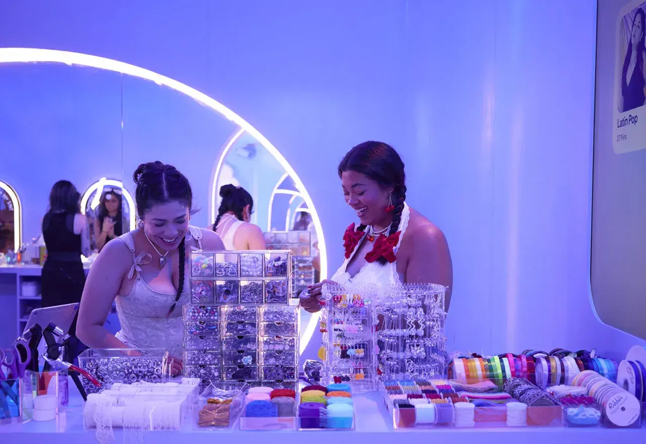 Dos mujeres sonríen en un puesto de accesorios para el cabello y cintas, parte de eventos de marca.