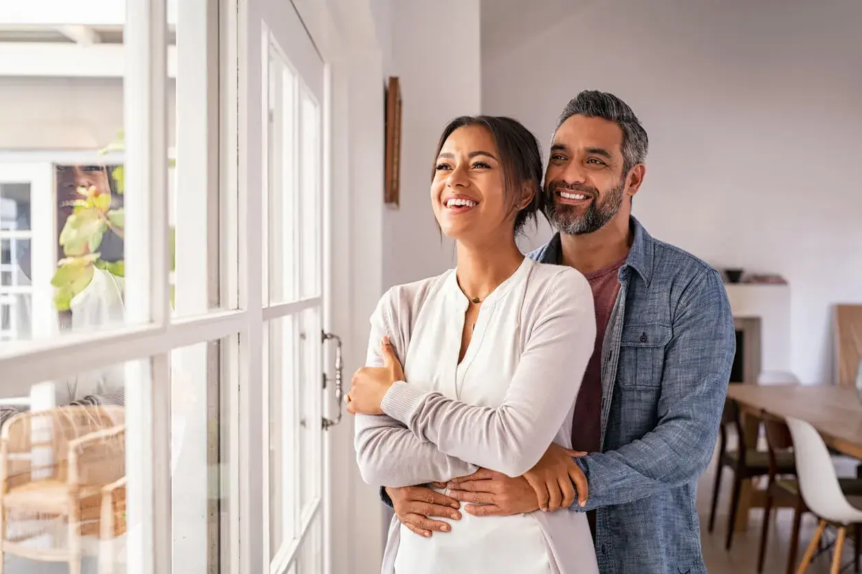 Pareja sonriente y abrazada, mirando por la ventana. Refleja cómo debe ser una relación de pareja: amor, apoyo y futuro compartido.
