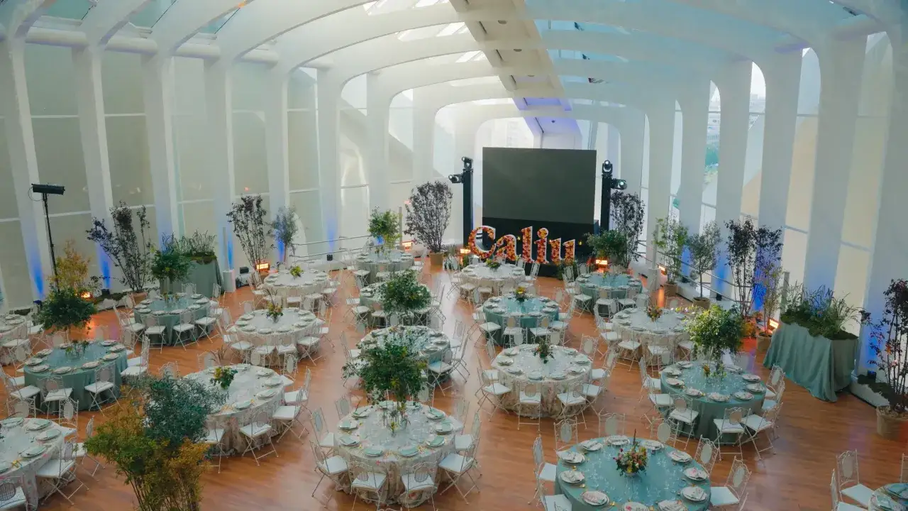 Salón moderno decorado para eventos empresariales, con mesas redondas vestidas y sillas blancas. Al fondo, un gran cartel floral con el nombre 