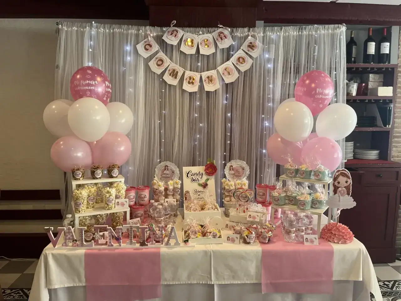 Mesa de dulces para fiestas sorpresa de comuni&oacute;n, con globos rosas y blancos, banderines y nombre 