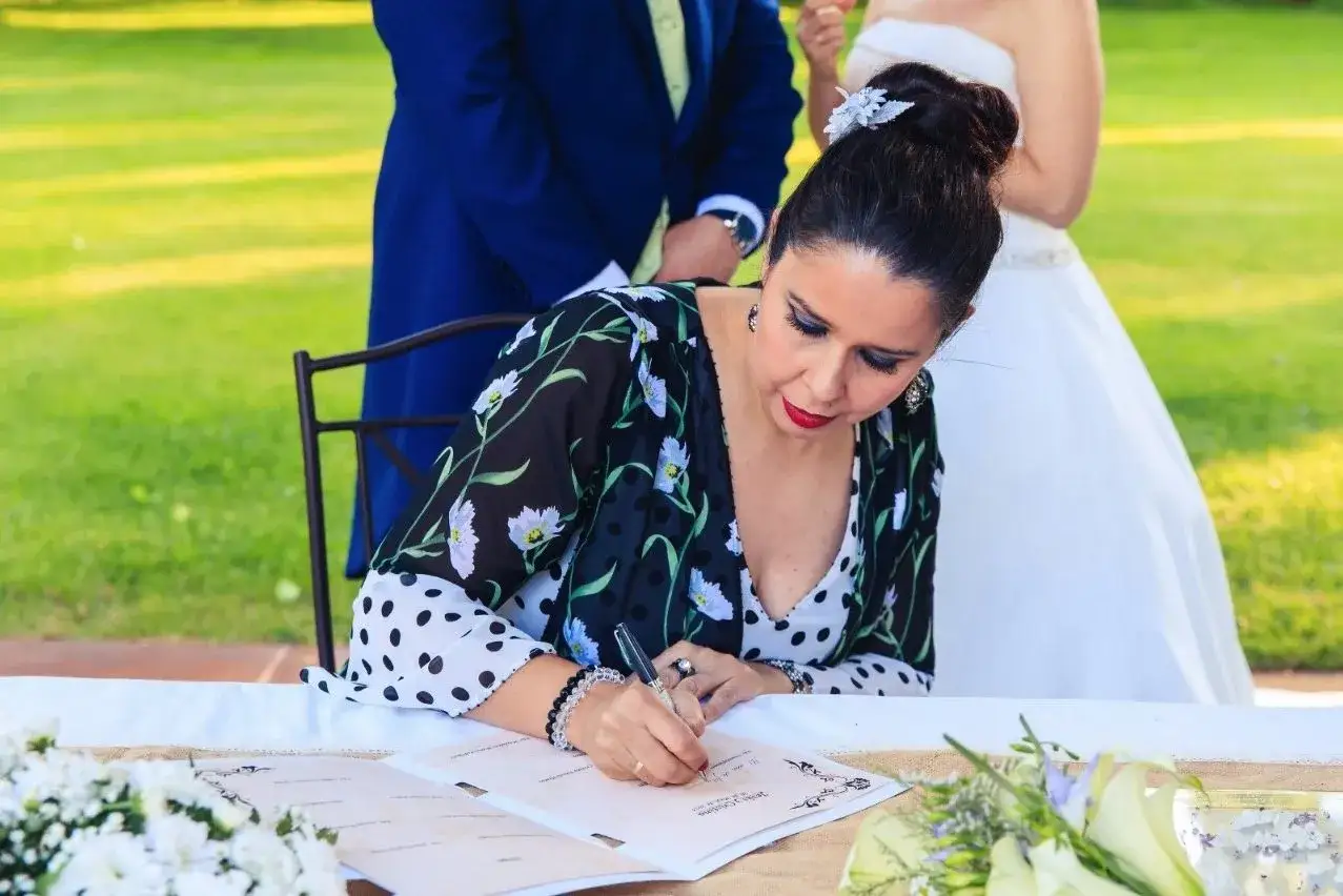 Una mujer con un vestido estampado firma un documento para oficializar una boda civil. Detr&aacute;s, se ven el novio y la novia.
