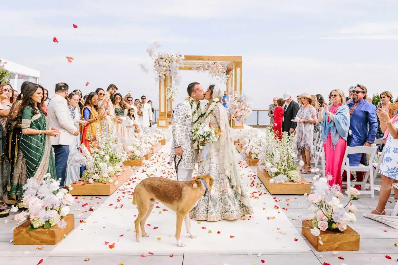 Una pareja se besa en su boda simbólica al aire libre, rodeada de invitados y flores. Un perro observa la escena.
