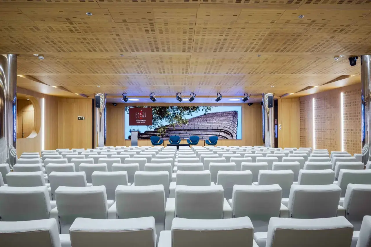 Sala de conferencias moderna, lista para un congreso evento. Butacas blancas, pantalla gigante con logo y arquitectura, y tres sillas azules en el escenario.