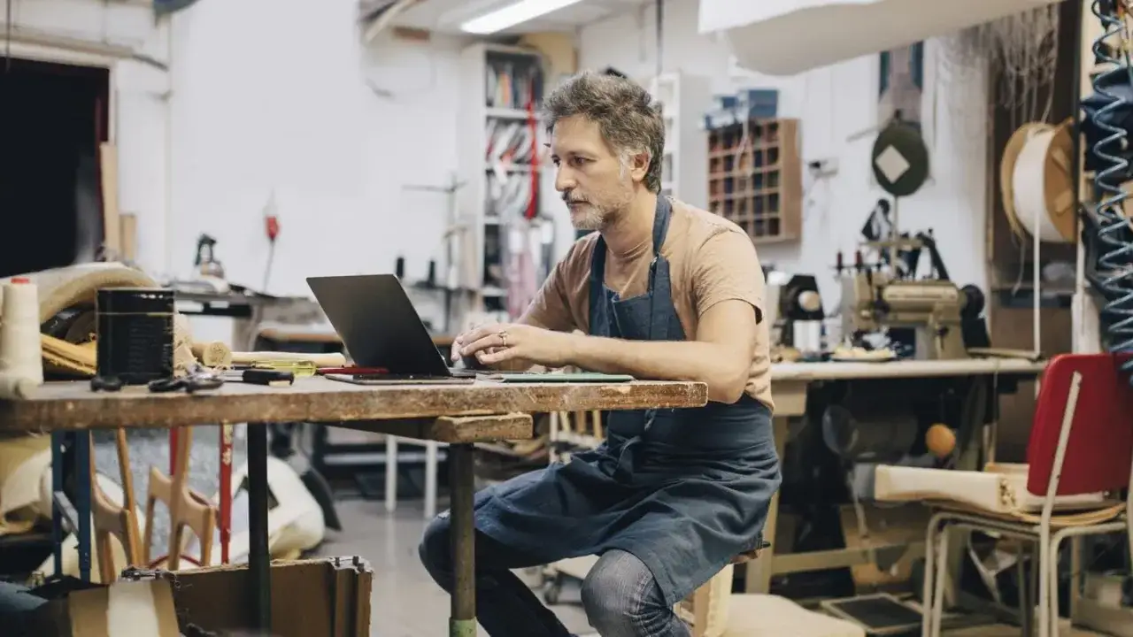 Hombre con delantal trabajando en su taller, gestionando su negocio online. Podría ser un falso autónomo si no tiene control real sobre su trabajo.