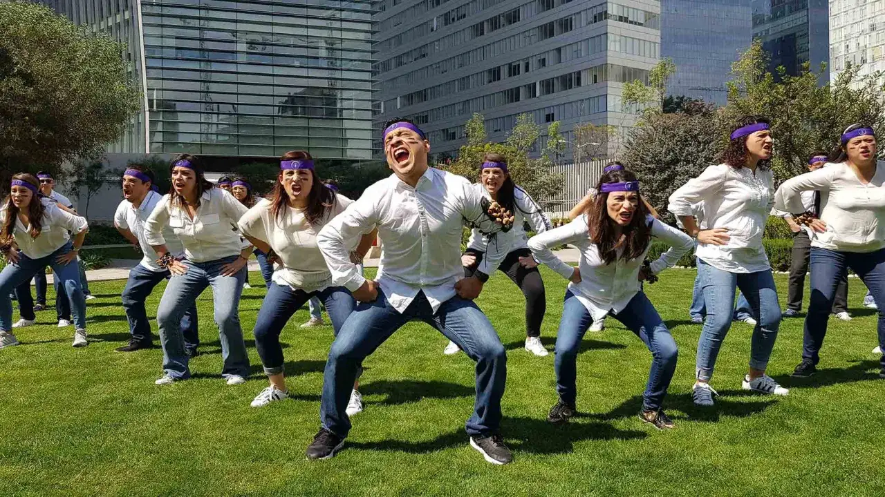 Grupo de personas en actividades para empresas team building, con vinchas p&uacute;rpuras y ropa blanca, bailando en un c&eacute;sped frente a edificios modernos.
