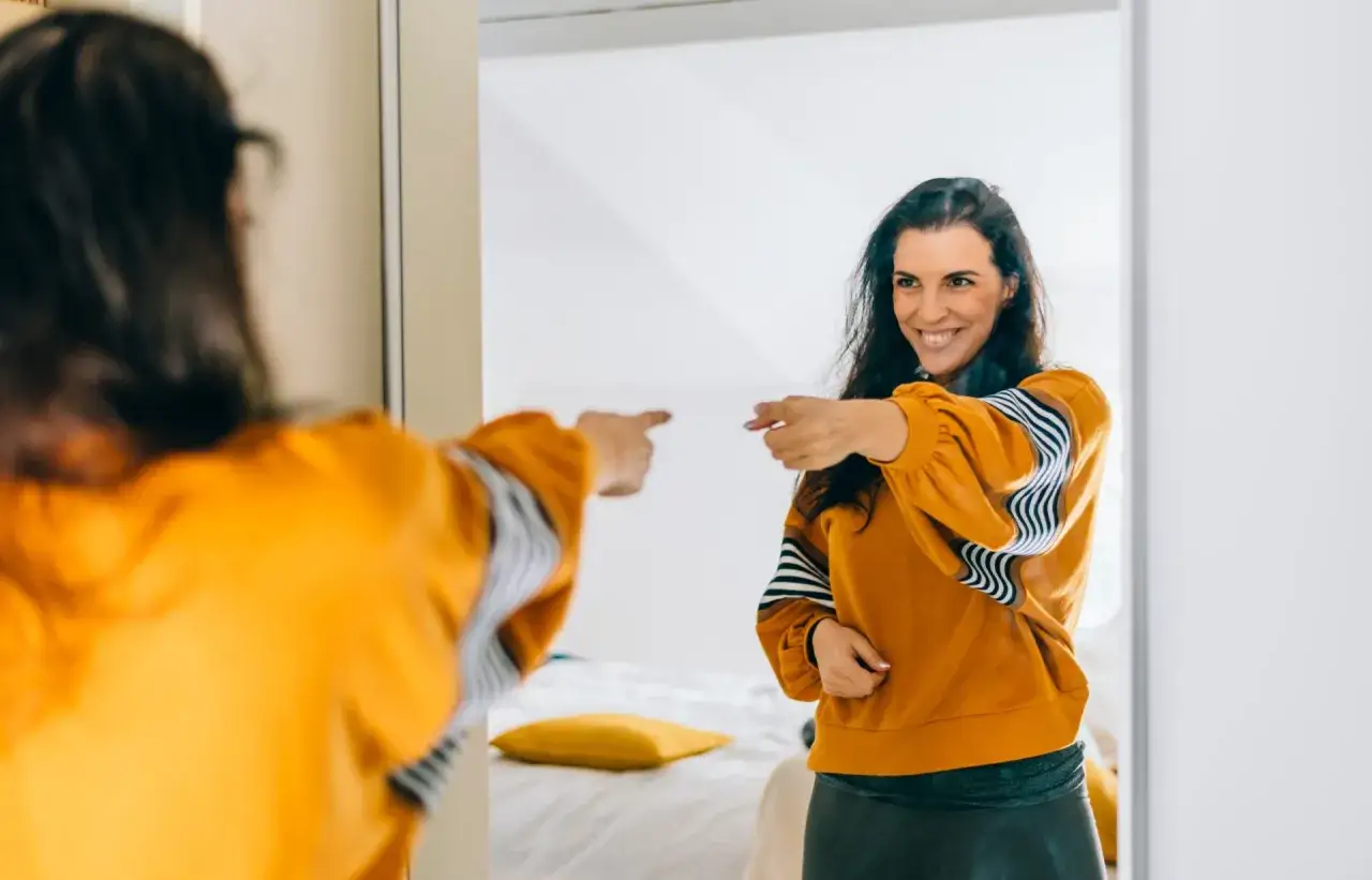 Una persona insegura puede cambiar. Mujer sonriente se se&ntilde;ala a s&iacute; misma en el espejo, con confianza.
