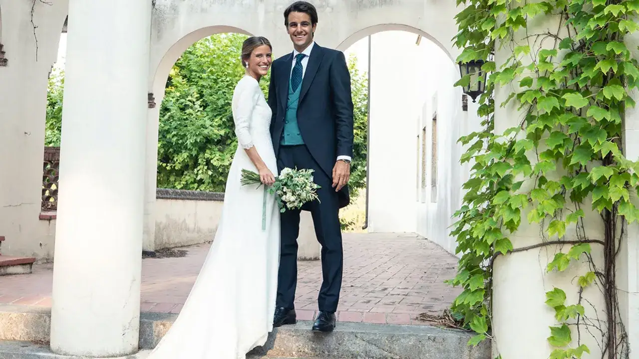 Novios sonriendo, ella con vestido blanco y ramo, él con traje oscuro. Un escenario perfecto para bodas vintage.