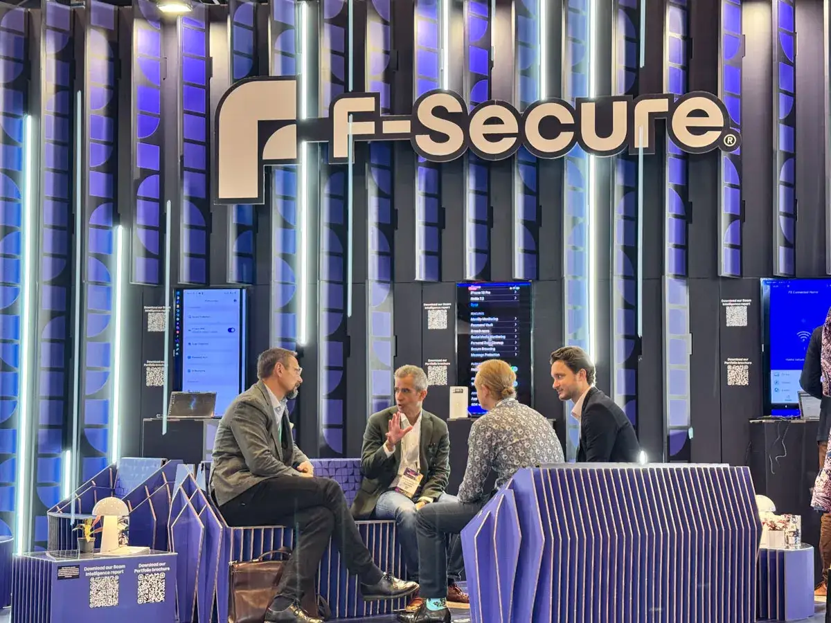 Hombres conversan en un stand de F-Secure durante ferias empresariales. El logo de la empresa destaca en un fondo azul y blanco.