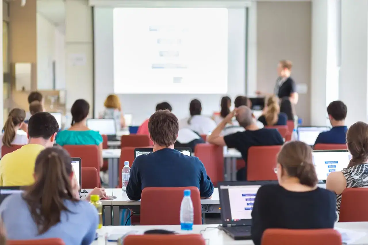 Asistentes en una sala de formaci&oacute;n presencial, atentos a la presentaci&oacute;n en pantalla y sus ordenadores port&aacute;tiles.