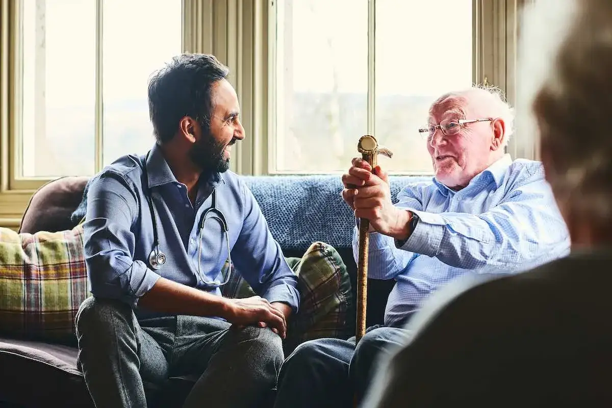 Médico y paciente mayor conversan con asertividad positiva. El doctor, con estetoscopio, escucha atentamente al anciano que se apoya en su bastón.